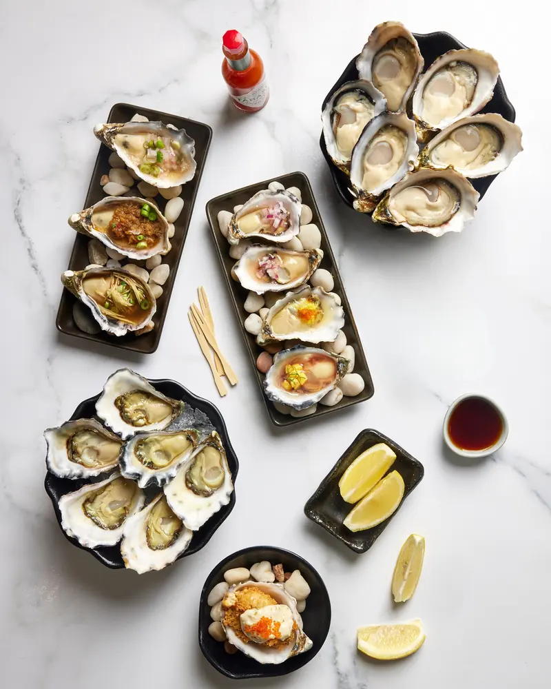 Dressed oysters on black trays of river stones, with natural oysters, lemon and dipping sauce on white marble